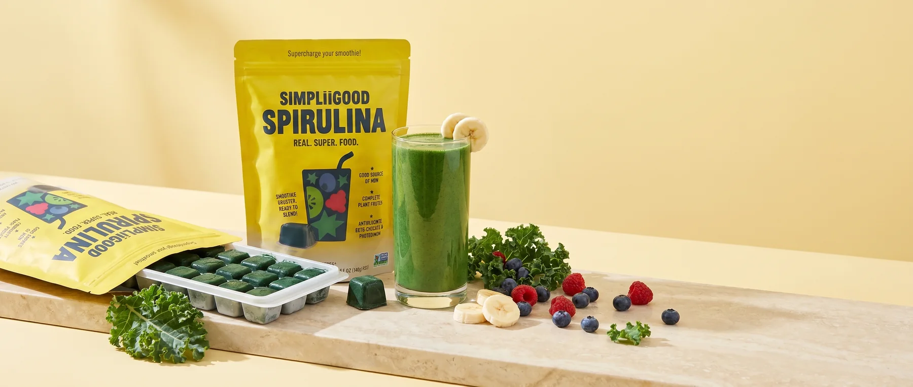 SimpliiGood spirulina pack with frozen cubes, a green smoothie and fresh fruit across a stone surface